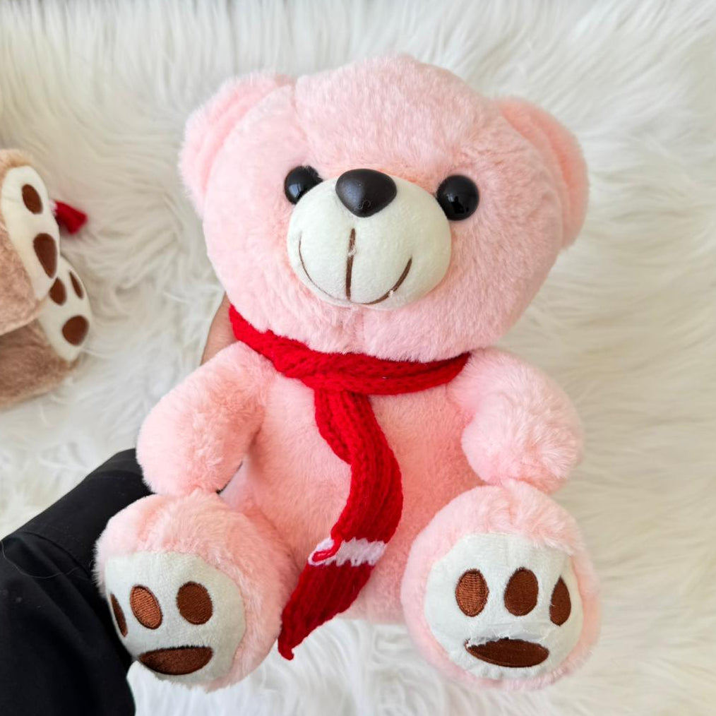 Pink teddy bear with a red scarf held by a person on a white fluffy surface.