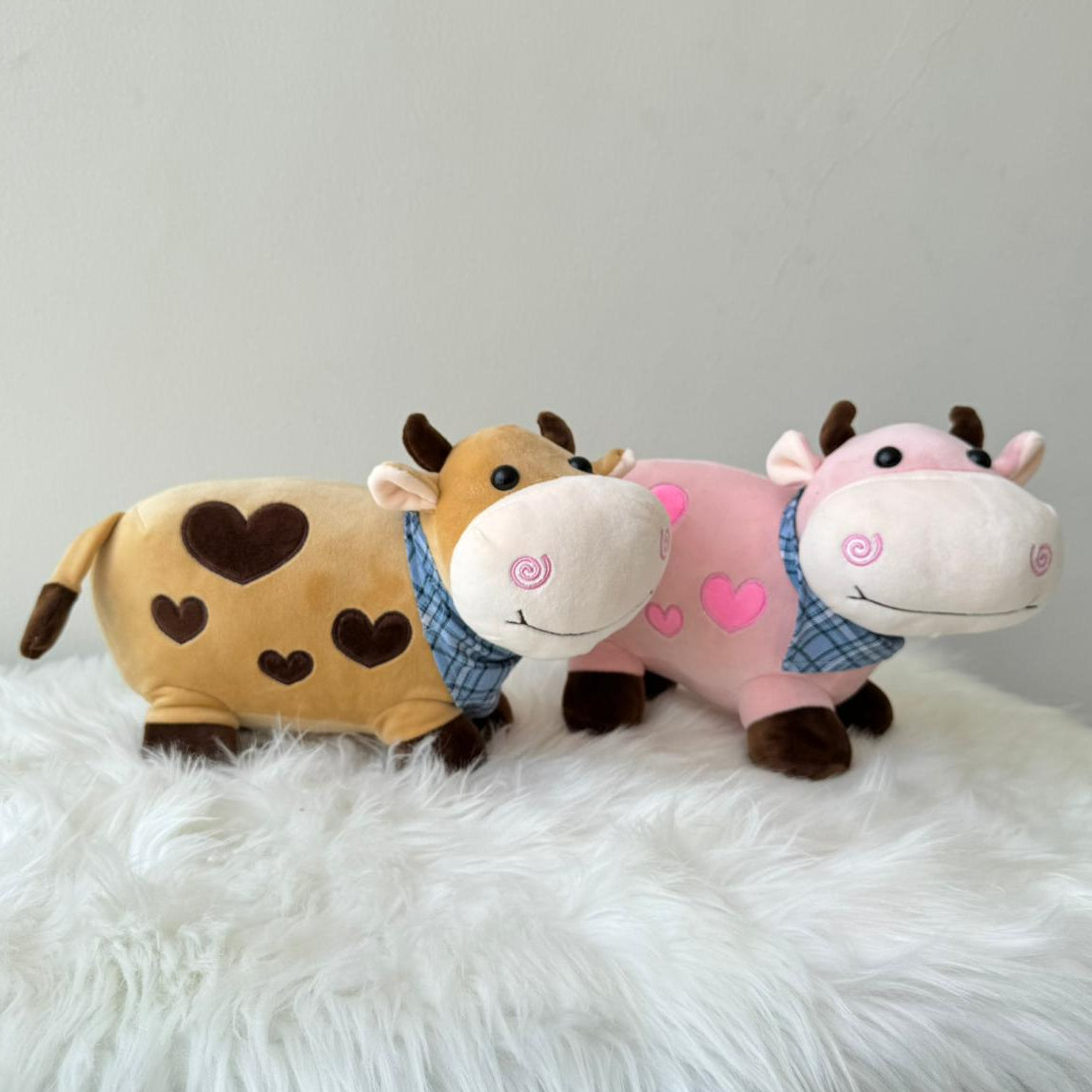 Two plush cow toys, one brown with heart patterns and one pink with heart patterns, on a white fluffy surface.