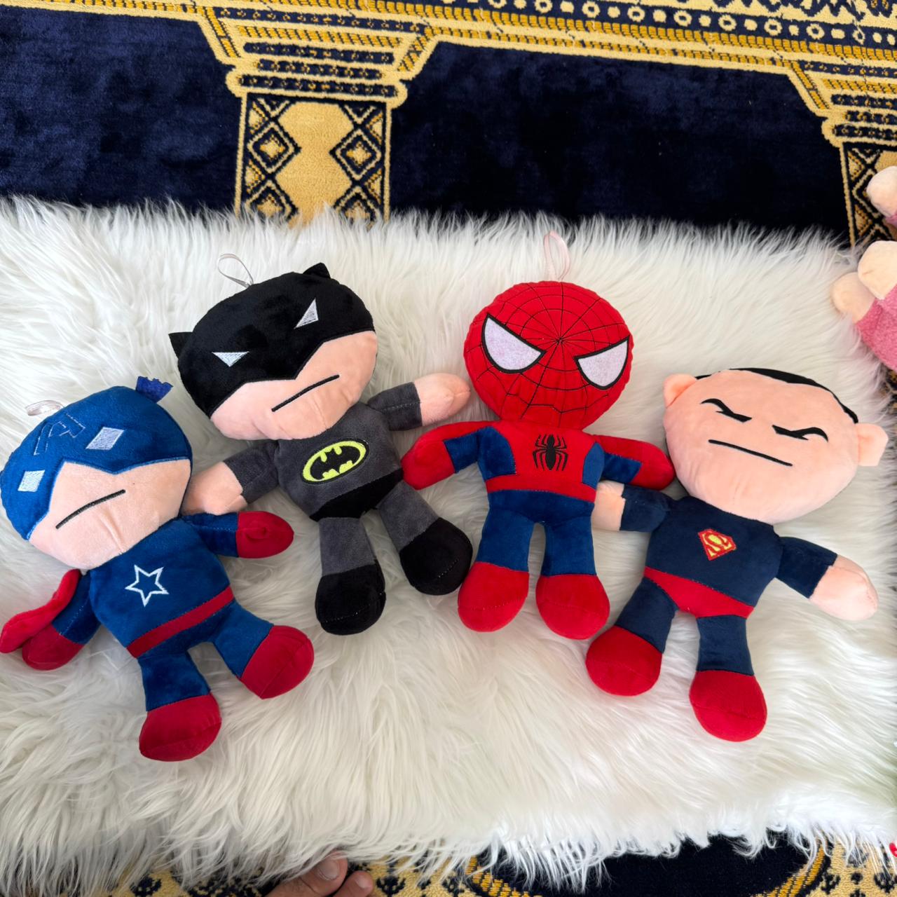 Four superhero plush toys on a fluffy white surface with a patterned rug in the background.
