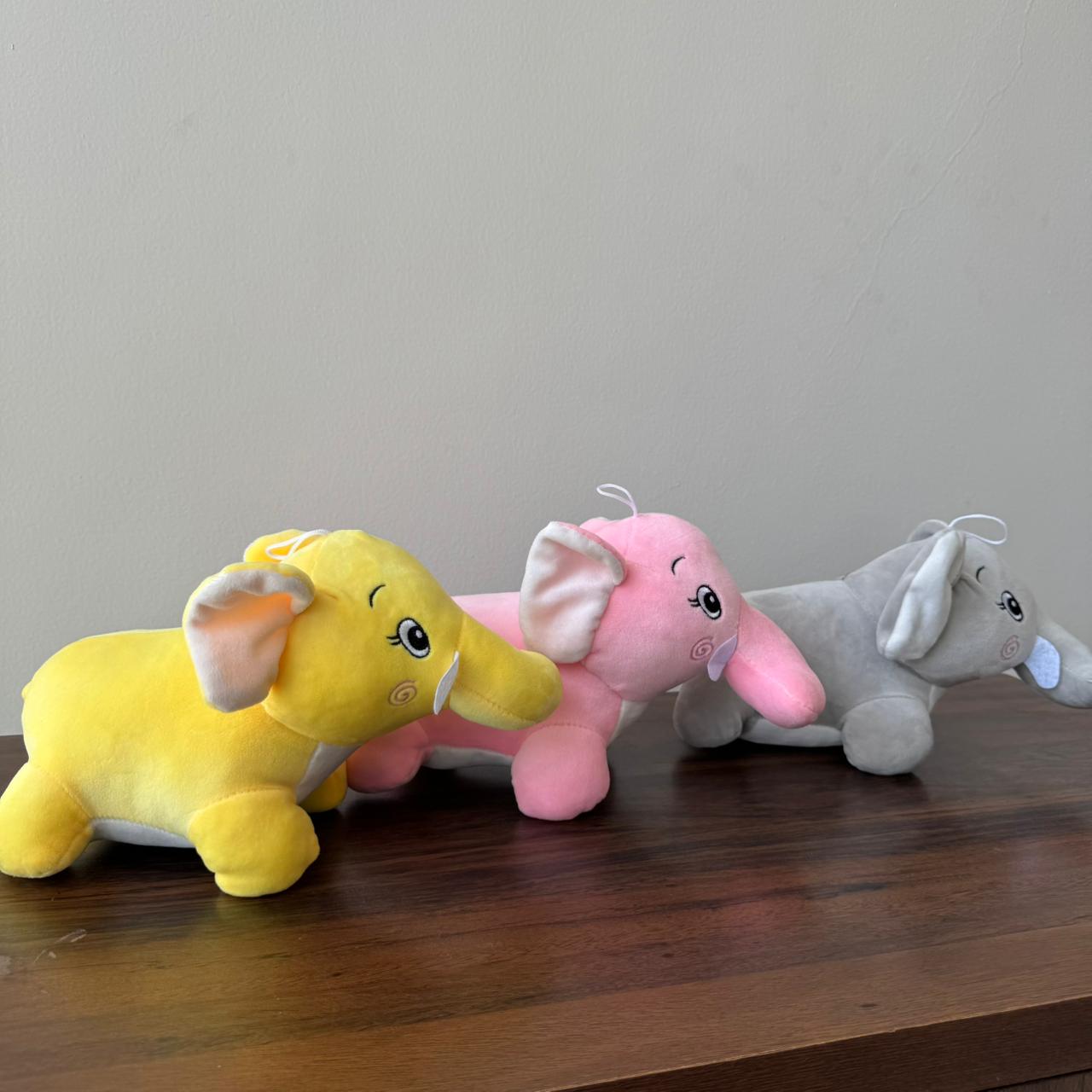 Three colorful elephant plush toys on a wooden surface with a plain background